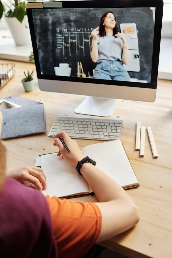 Student attending virtual class on a computer, taking notes at home.